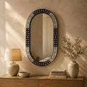 Oval wall mirror with black and white bone inlay and ornate frame, styled above wooden console table with table lamp, decorative vases, dried plants, and books