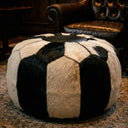 A round pouf ottoman upholstered in black and off-white cowhide panels, styled in a living room setting on a patterned carpet with a leather armchair in the background.