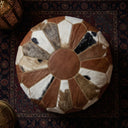 A round pouf ottoman made of patchwork animal-hide panels in brown, white, black, and gray, with a solid brown center and decorative gold-toned stitching, shown from a top-down view on a dark patterned rug.