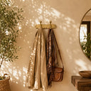 A brass double coat rack hanging on a warm beige wall, holding a light beige jacket and a brown patterned scarf, with a brown leather shoulder bag draped from the rack. Sunlight with leaf shadows is cast across the wall, and a potted plant and wooden table are visible in the room.