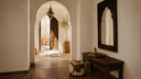 Moroccan-style interior hallway with arched doorway, dark wood console table, ornate wall mirror, hanging brass lantern, and patterned tile floor.