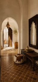A warm, Moroccan rustic interior hallway with terracotta patterned tile flooring, a dark wooden console table, a large arched mirror, and a round cowhide pouf.