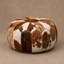 Round cowhide pouf ottoman with a patchwork pattern of brown, black, and white hair-on-hide panels, set against a neutral beige background.
