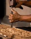 Close-up of a person's hands holding a wooden mallet and metal chisel to carve or shape a wooden panel, with wood shavings scattered on the workbench.