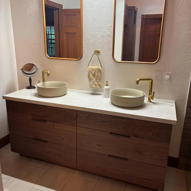 A double-sink bathroom vanity with a dark wood cabinet, white countertop, round vessel sinks, and gold faucets, shown in a real bathroom setting with mirrors and wall outlets visible.