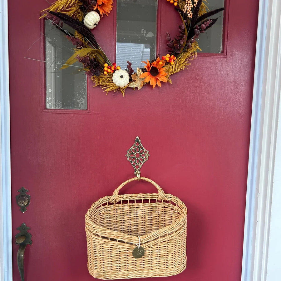 A natural woven wicker basket with a top handle hangs from an ornate metal hook on a dark red front door. Above the basket, a fall-themed wreath with orange sunflowers, small pumpkins, and autumn leaves decorates the door.