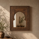 An ornate rectangular wall mirror with a carved dark brown wooden frame, mounted on a textured beige wall. The mirror features a central arched opening and intricate geometric patterns around the border.