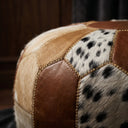 Close-up of a patchwork pouf or ottoman featuring brown leather panels, tan cowhide, and black-and-white dalmatian-patterned hide sections, with gold-toned stitching.
